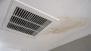 Realistic upward view of a white home ceiling with a modern air conditioning vent beside a spreading water stain and a single droplet forming on the drywall.